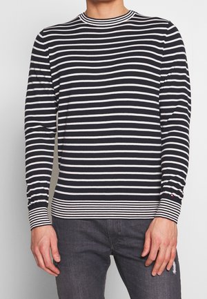 Mand iført sort-hvidt langærmet skjorte med horisontale striber, ensfarvede manchetter og kant, kombineret med mørkegrå jeans.
