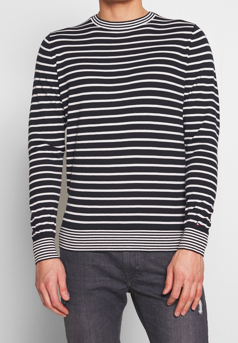 Homme portant une chemise à manches longues rayée horizontalement noir et blanc avec des poignets et un bas unis, associée à un jean gris foncé.