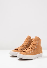 Tan high-top sneakers made of suede with white speckled rubber soles. Features circular metal eyelets and flat laces. Smooth texture.