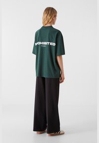Donkergroen oversized t-shirt met witte tekst "PROHIBITED 10173, BERLIJN" op de achterkant, gecombineerd met wijde zwarte broek en casual sneakers.