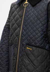 Πουπουλένιο μπουφάν από νάιλον σε μαύρο και ναυτικό μπλε. Διαθέτει χρυσό �φερμουάρ, γιακά από κορντούρα και τσέπη με το κέντημα "Barbour" σε χρυσό χρώμα.