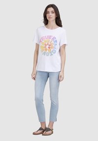 T-shirt en coton blanc décontracté avec un texte graphique circulaire multicolore. Associé à un jean skinny bleu clair délavé et des sandales marron ornées.