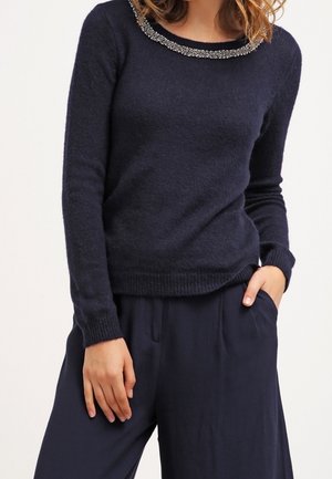 Femme portant un pull à manches longues bleu marine foncé avec un col décoratif orné de perles et un pantalon ample assorti, une main dans la poche.