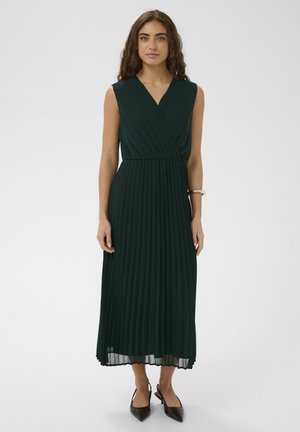 Žena s kovrčavom kosom u bezrukavnoj tamnozelenoj plisiranoj midi haljini i crnim špicastim sandalama s remenčićem, stoji na bijeloj pozadini.