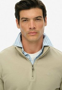 Beżowy sweter zapinany na zamek, wykonany z fakturowanej tkaniny, noszony na jasnoniebieskiej koszuli w paski. Widoczne logo na klatce piersiowej.