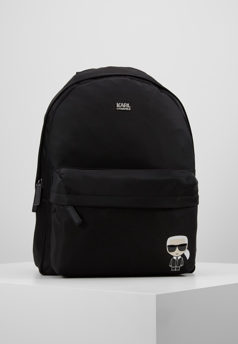 Sac à dos noir avec poche zippée à l'avant, logo Karl Lagerfeld en haut et petite figurine cartoon Karl Lagerfeld sur la poche inférieure droite.