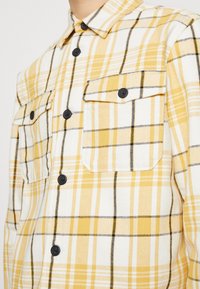 Chemise en flanelle à carreaux jaunes et blancs avec des accents noirs. Elle possède deux poches poitrine et des boutons noirs. Texture douce, col à boutons.