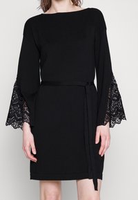 Černé šaty s žebrovaným výstřihem, dlouhými zvonovými rukávy s komplikovanou černou krajkou a pasem se sponou v odpovídající barvě. Hladká textura látky.