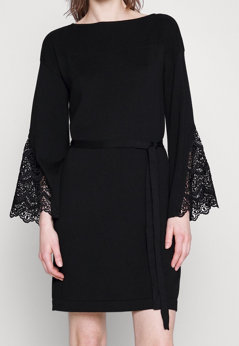 Černé šaty s žebrovaným výstřihem, dlouhými zvonovými rukávy s komplikovanou černou krajkou a pasem se sponou v odpovídající barvě. Hladká textura látky.