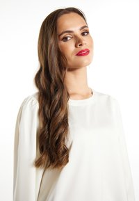 Bílá dlouhá halenka s hladkou texturou. Vlnité, dlouhé hnědé vlasy. Červená rtěnka. Jednoduchý design, bez viditelných vzorů nebo akcentů.