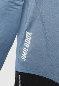 Hellblaues Sportshirt mit strukturiertem Muster, taillierter Passform und weißem Logo, das auf der linken Seite aufgedruckt ist. Sichtbare Nähdetails.