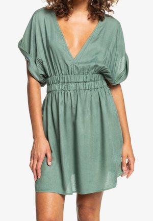 Kobieta w krótkiej, luźnej zielonej sukience z głębokim dekoltem w kształcie litery V i marszczonym, elastycznym pasem, stojąca na białym tle.