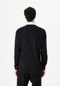 Svart langermet skjorte laget av glatt stoff, med hvit krage, knappmansjetter og rett kant. Bakoversikt vist.