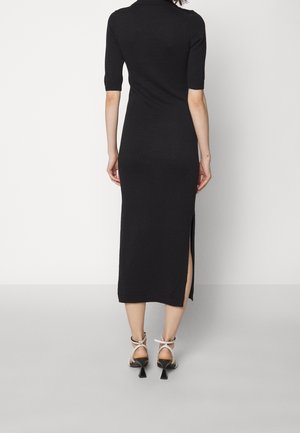 Femme debout de dos, portant une robe midi noire avec une fente latérale et des sandales blanches à talons hauts.
