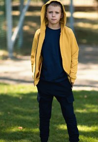 Żółta bluza z kapturem zapinana na zamek nałożona na granatową koszulkę z okrągłym dekoltem, w połączeniu z granatowymi joggerami cargo. Gładka tekstura o luźnym kroju.