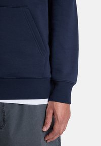 Sudadera azul marino con un bolsillo tipo canguro, puños y dobladillo de canalé. Textura suave con un diseño ajustado, llevada sobre una camisa blanca.