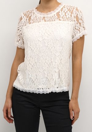 Kobieta ubrana w białą, kwiecistą koronkową bluzkę z krótkim rękawem i ozdobnymi falbankami, w połączeniu z czarnymi spodniami, na jednolitym tle.