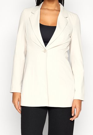 Personne portant un blazer beige clair à un bouton sur un haut noir et un pantalon noir, debout avec les mains détendues le long du corps.