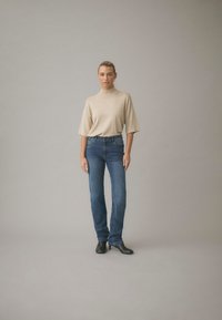 Kvinna med tillbakadraget hår som bär en beige kortärmad polotröja, blå jeans och svarta stövletter med klack mot en enkel grå bakgrund.