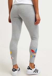 Grå leggings gjorda av elastiskt material med flerfärgade Adidas-logotillbehör i gult, rött och blått vid anklarna. Stylat med svarta sneakers.