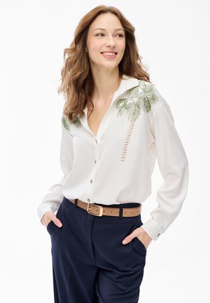 Uśmiechnięta kobieta o falujących włosach nosi białą bluzkę z haftem palmy, wpuszczoną w granatowe spodnie z jasnobrązowym paskiem, z rękami w kieszeniach.
