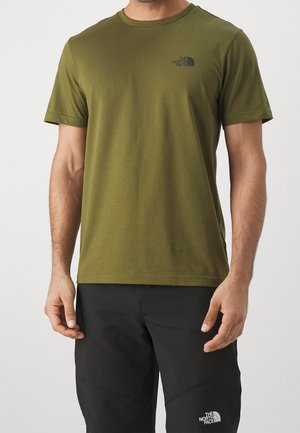 Mężczyzna w oliwkowej koszulce z krótkim rękawem i czarnych spodniach, obie z logo The North Face na lewej piersi i udzie.