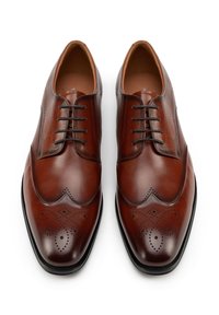 Paar aus poliertem braunem Leder gefertigte Herrenschuhe mit dekorativen Perforationen und schwarzen Schnürsenkeln, von oben auf weißem Hintergrund gesehen.