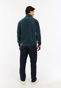 Chaqueta de forro polar verde oscuro con cuello alto, mangas largas y puños acanalados, combinada con jeans de mezclilla azules y zapatillas blancas.