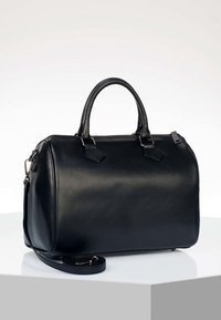 Sac à main en cuir noir avec deux poignées, fermeture éclair et bandoulière amovible, posé sur une surface blanche.