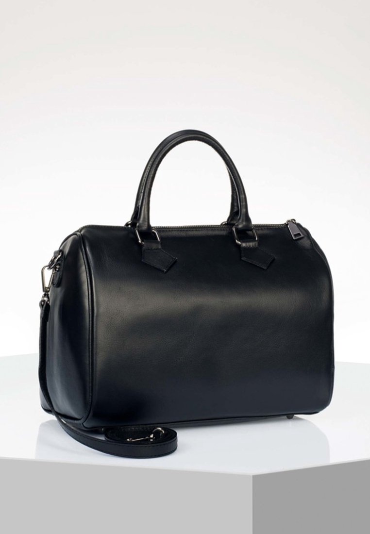 Sac à main en cuir noir avec deux poignées, fermeture éclair et bandoulière amovible, posé sur une surface blanche.