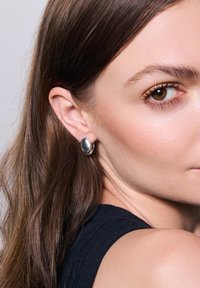 Srebrne kolczyki w kształcie owalu, z gładką, odblaskową powierzchnią. Noszone na uchu modelki, podkreślające ich szczegóły wykonania.