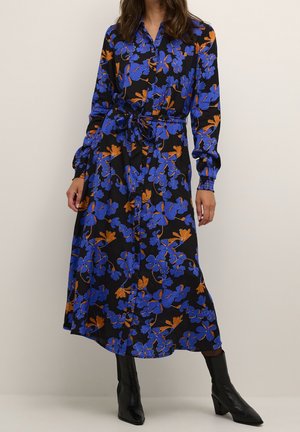 Kvinde iført en sort midi-kjole med lange ærmer, blå og orange blomsterprint, bælte i taljen og sorte ankelstøvler med hæl.