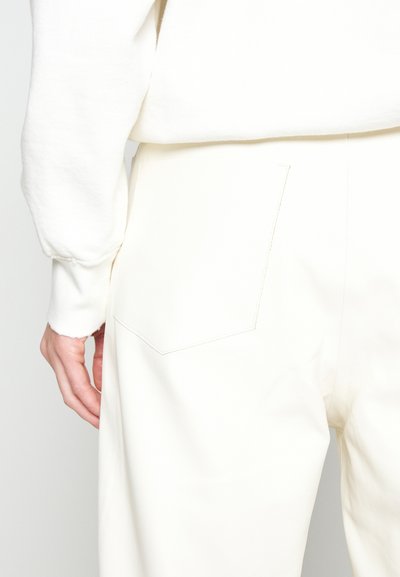 Άτομο που φοράει λευκό παντελόνι και λευκή μπλούζα με μακριά μανίκια, δείχνοντας το αριστερό χέρι και με ορατή πίσω τσέπη στο παντελόνι.