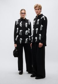 Τα μαύρα πουκάμισα έχουν λευκές υφασμάτινες αποκοπές που μοιάζουν με κορδέλες, συνδυασμένα με φαρδιά μαύρα παντελόνια και μαύρο τσάντα. Το υφασμάτινο υλικό έχει έντονη υφή που δημιουργεί αντίθεση.