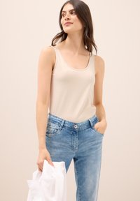 Canotta beige senza maniche con scollatura a girocollo, abbinata a jeans azzurri chiari. I jeans presentano un bottone e tasche, indossati con una camicia bianca.