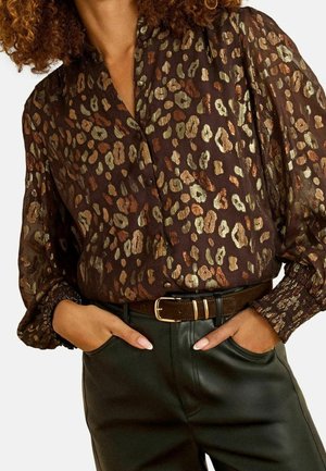 Femme portant un chemisier marron métallisé à imprimé léopard avec des manches bouffantes et un pantalon noir taille haute avec une ceinture marron. Mains dans les poches.