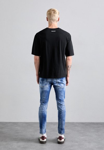 Zwarte T-shirt met korte mouwen, gecombineerd met vervaagde blauwe spijkerbroeken met witte verfspatten. Sneakers met witte zolen en kleurrijke accenten.