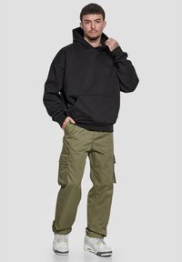 Svart hettegenser med frontlomme, kombinert med olivengrønne cargo-bukser med sidelommer, og hvite joggesko med grå detaljer.