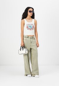 Woman wearing graphic tank top, oversized light-wash jeans, white sneakers, sunglasses, holding small white handbag with black handles.