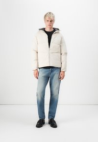 Chaqueta acolchada blanca con capucha, que presenta un cierre de cremallera y detalles cosidos. La lleva puesta sobre una camiseta negra y la combina con unos jeans azules.
