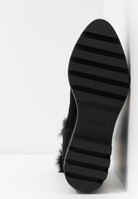 Semelle noire d'une chaussure DKNY avec des crampons horizontaux épais, inclinée contre un mur blanc et un sol blanc.