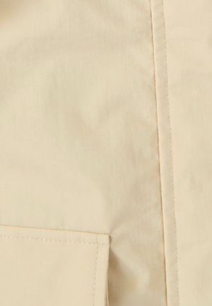 Beiger, leichter Stoff mit einer glatten Textur, ausgestattet mit einer seitlichen Tasche und dezenten Steppnähten entlang der Naht.