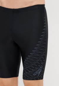 Calções de banho pretos feitos de um tecido elástico, com um padrão texturizado de zigue-zague em azul claro no lado esquerdo e o logótipo Speedo perto da bainha.