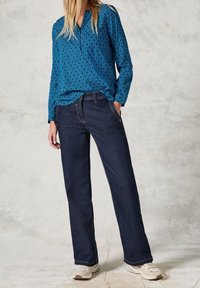 Blouse à manches longues à motifs bleus avec un col en V, associée à un pantalon droit en denim foncé et des baskets claires, avec des coutures et des poches visibles.