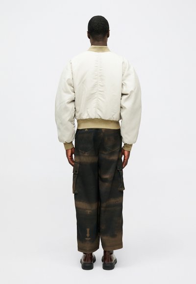 Jachetă bomber bej deschis cu mâneci pufose, asortată cu pantaloni cargo negri cu un model gradient și buzunare laterale. Pantofi maro.