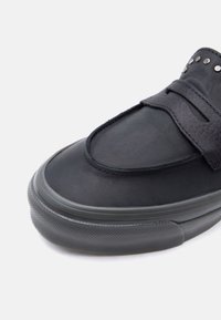 Gros plan sur un mocassin noir avec une semelle épaisse en caoutchouc et des clous métalliques décoratifs sur la bride supérieure.