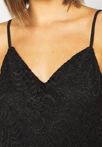 Camisole en dentelle noire avec un col en V et de fines bretelles, présentant des motifs floraux complexes et une finition texturée.