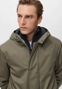 Chaqueta impermeable verde olivo con capucha, cierre frontal con cremallera, dos capas de tejido y botones a presión. Presenta una textura suave y un diseño informal.