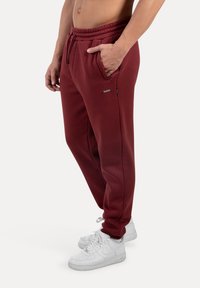 Muž v tmavě červených teplácích se elastickým pasem a šňůrkou, v bílých teniskách, stojící s jednou rukou v kapse.