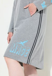 Abito in cotone grigio con tasca laterale, testo grafico blu "MAGE" e design di un uccello, con strisce bianche e nere lungo i lati.